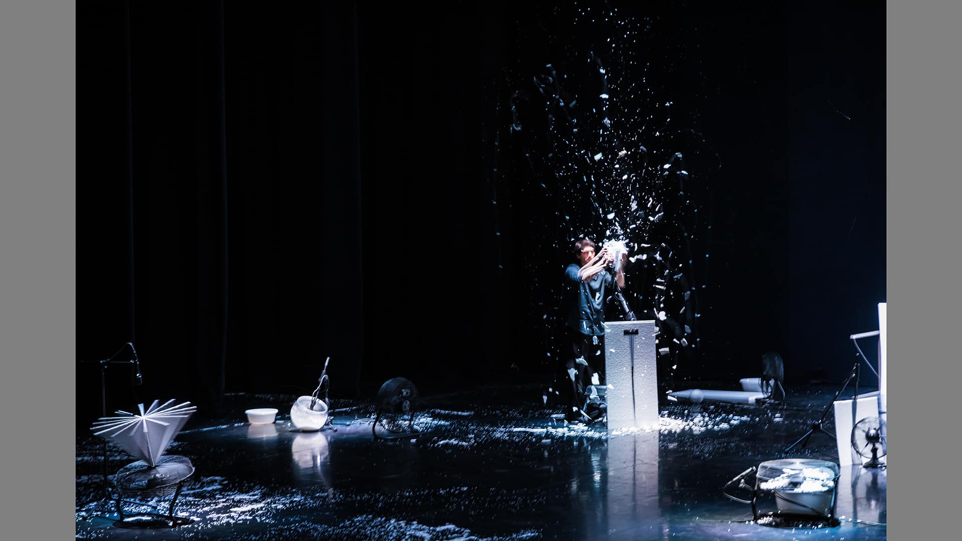 A photo of the show in progress, featuring a wide shot of the black stage space, scattered with small white objects and with a small human figure half-lit with white, holding a ball in a snowstorm