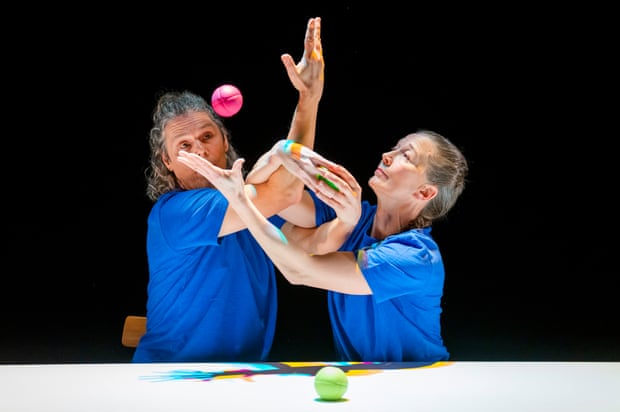 Photo shows the upper bodies of a man and a woman sat at a table. They wear matching blue t-shirts and their arms are comlicatedly intertwined and in motion, passing two coloured balls between them