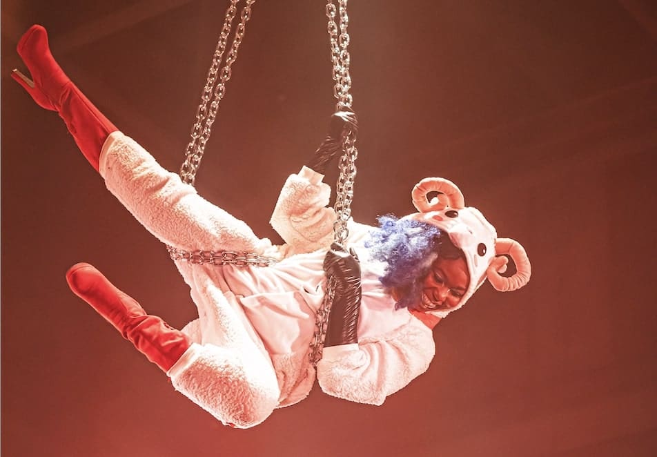 Photo of a smiling black woman with blue hair holding herself horizontally on suspended metal chains. She is wearing a fluffy white sheep onesie with curly horns, and long red high-heeeled boots.
