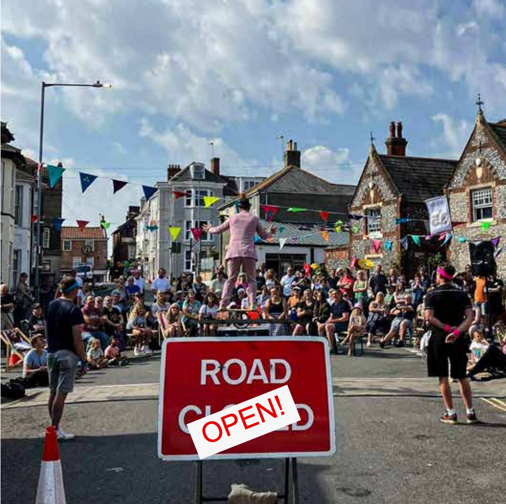 Photo of an English street hung with bunting. A man in a pink suit stands with his back to the camera on top of a rola-bola - a plank of wood balanced on top of a football sized cylinder. Smiling rows of audience members are sat on deckchairs or standing behind them watching him. Much closer to the camera is a red street sign that says Road Closed and a traffic cone. A digital edit has added a layer over the photo that covers up the word Closed with the word Open.